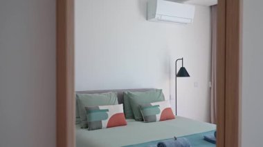 Rahat yatak odasında yumuşak yastıklar, katlanmış havlular, şık bir zemin lambası ve temiz beyaz bir duvarda gizli bir havalandırma ünitesi olan büyük bir yatak var.