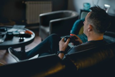 Kanepede oturan ve elinde joystick tutan yetişkin bir adam konsolda video oyunu oynuyor.
