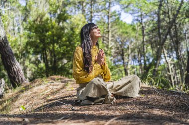 Elleri sımsıkı bağlı meditasyon yapan bir kadın ormanda gökyüzüne bakıyor.