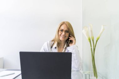 Gülümseyen kadın doktor, bir hastayla telefonda konuşurken tıp bürosundaki kameraya bakıyor..