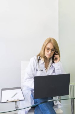Tıbbi ofisindeki dizüstü bilgisayarında bir hastanın tıbbi kayıtlarına bakarken telefonda konuşan bir kadın doktor..