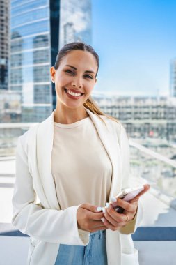 Girişimci bir kadın kameraya bakarken gülümsüyor ve cep telefonu tutuyor. Beyaz bir ceket ve kot pantolon giyiyor. Girişimcilik ve modernlik kavramı.