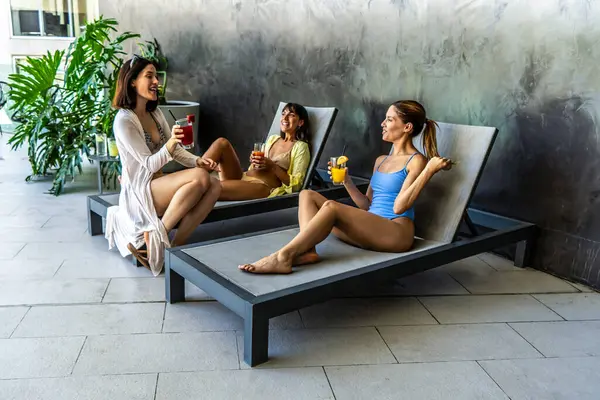 Üç kadın şezlongda oturmuş meyve suyu içiyor. Gülümsüyorlar ve birbirlerinin arkadaşlığından zevk alıyorlar.