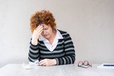 Orta yaşlı kızıl saçlı bir kadın bir elini alnında tutuyor..