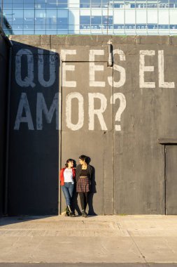 Young lesbian couple gazing into each other's eyes, leaning against a black wall with graffiti that reads 