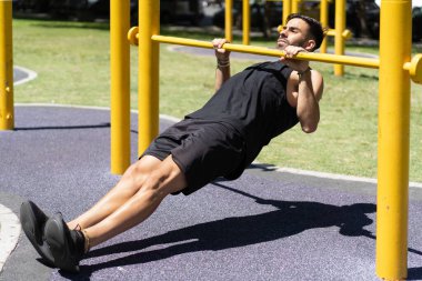 Fit young man executing inverted rows on outdoor gym bars. clipart