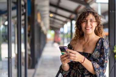 Mid-adult curly woman using her smartphone while looking at the camera in a modern shopping district clipart