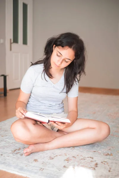 Cute young girl reading paper book while sitting on carpet at home