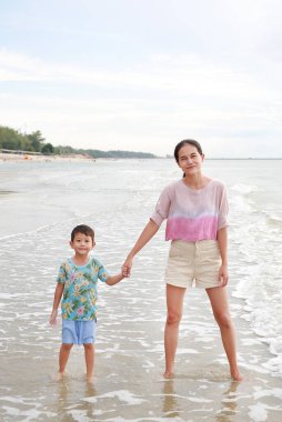 Asyalı anne ve küçük çocuk tropikal kumsalda duruyorlar. Mutlu aile anne ve oğlu yaz tatilinde eğleniyor..