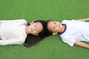Asyalı küçük oğlan kameraya bakıyor ve kız çocuk yeşil çimlerin üzerinde yatarken gözlerini kapatıyor. Görünümün üstünde.