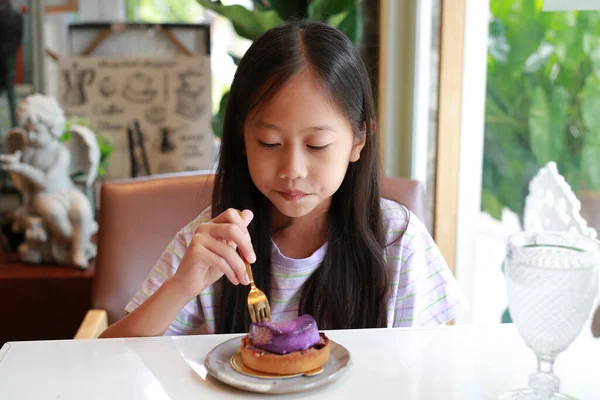 stock image Asian girl child sitting by table and eating Blueberry Cheesecake cake in Cafe. Kid eats tasty food.