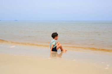 Asyalı küçük çocuk tropikal kumsalda oturmuş denize bakıyor..