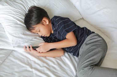 Küçük Asyalı bir çocuk akıllı telefon kullanıyor ve telefonu yatakta tutarken uyuyakalıyor..