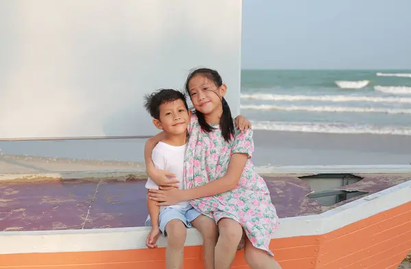 幸せなアジアの男の子と少女の肖像画は セーリングボートに座って 夏の休日にビーチでカメラを見ながら座って抱き合っています ストック画像