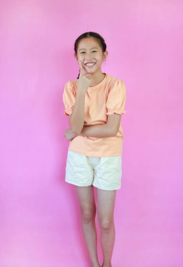 Neşeli Asyalı kız çocuğu parmaklarını yanağında tutuyor. Çocuk ayakta ve kameraya bakıyor. Pembe stüdyo arka planında izole edilmiş..