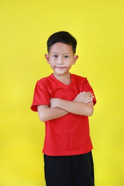 Gülümseyen Asyalı çocuk göğsünün üzerinde durup kollarını kavuşturarak sarı arka planda izole edilmiş kameraya bakıyor..