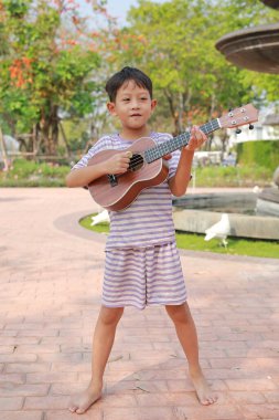 Asyalı küçük çocuk yaz bahçesinde dikilirken klasik ukulele çalıyor..