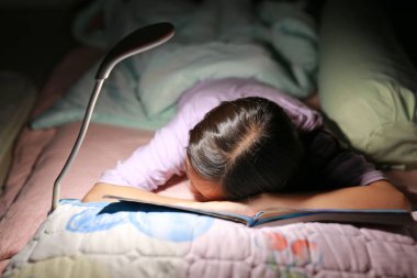 Genç Asyalı kız geceleri yatağında lamba ışığı olan bir kitap okurken uyuyor. Yakın çekim ve yumuşak odaklanma.