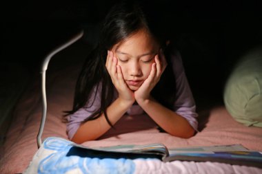 Asyalı genç bir kız gece yatakta yatarken lambalı bir kitap okuyor. Yakın çekim..