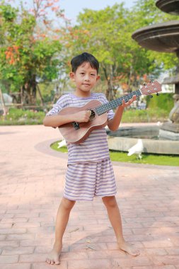 Yazlık bahçede dikilirken kamerayla klasik ukulele çalan Asyalı çocuğun portresi..