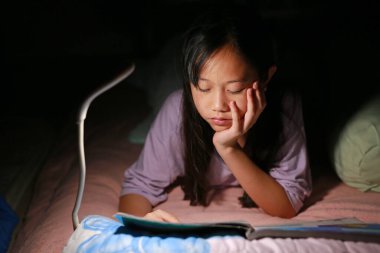 Asyalı genç bir kız gece yatakta yatarken lambalı bir kitap okuyor. Yakın çekim..