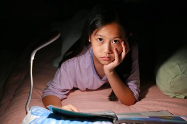 Asyalı genç bir kız gece yatakta yatarken lambalı bir kitap okuyor. Yakın çekim..