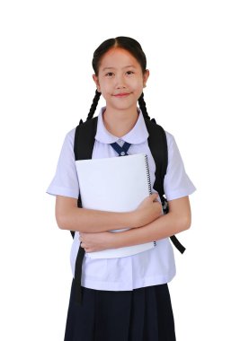 Okul üniformalı Asyalı kız beyaz stüdyo arka planında tek başına dururken sırt çantası taşıyıp bir kitaba sarılıyor..