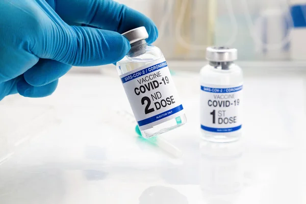 手を握るCovid 19ワクチン2回目の投与と背景1回目のワクチンでタグ付けされたワクチン用バイアル 医師はラベルにワクチンの2番目の用量の名前を持つコロナウイルスワクチン瓶を保持 — ストック写真