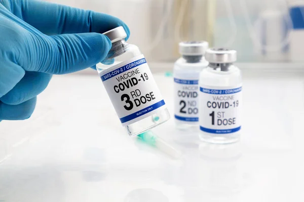 持用Covid 19疫苗Vial进行疫苗接种 标有第三剂和背景1 2剂疫苗 标签上印有第三剂疫苗名称的Coronavirus疫苗瓶的医生 — 图库照片