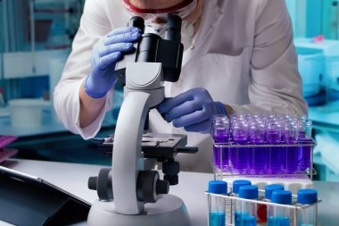 Mor sıvıyla çevrili test tüpleriyle çevrili bir araştırma laboratuvarında mikroskop kullanarak bilimsel analiz yapan bir araştırmacı. Laboratuvarda test tüpleriyle mikroskop kullanan bilim adamı.