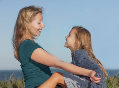 Neşeli anne portresi. Küçük kızı gülüyor ve mavi gökyüzü arkaplanıyla birbirine bakıyor.