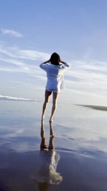 Bikini ve beyaz tişörtlü neşeli esmer kız okyanusun kenarında yürüyüş yapıyor.