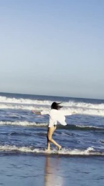 Bikini ve beyaz tişörtlü neşeli esmer kız okyanusun kenarında uçan martılarla koşan yaz akşamının tadını çıkarıyor.