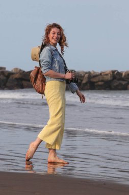Sırt çantası ve kamerasıyla sahilde yürüyen neşeli genç bir kadının portresi. 