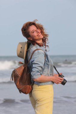 Sırt çantası ve kamerasıyla sahilde yürüyen neşeli genç bir kadının portresi. 