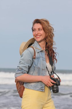 Sırt çantası ve kamerasıyla sahilde yürüyen neşeli genç bir kadının portresi. 