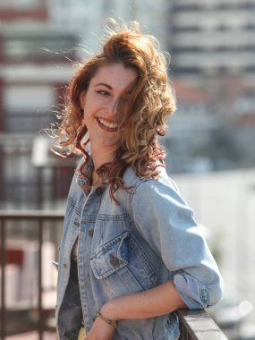 Arka planda şehirle birlikte balkonda duran neşeli kızıl saçlı genç kadının portresi.