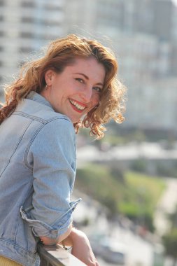 Arka planda şehirle birlikte balkonda duran neşeli kızıl saçlı genç kadının portresi.