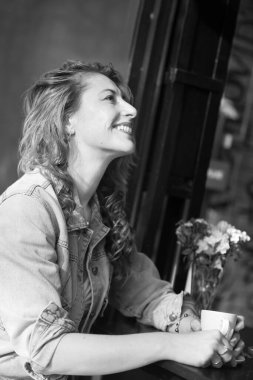 Açık hava kafeteryasındaki siyah beyaz masada oturan neşeli bir kadının portresi.