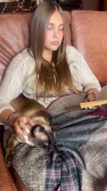 Kanepede kediyle kaplı bir şekilde oturan ve evde kitap okuyan genç bir kadın. 