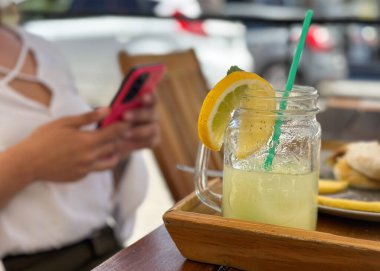 Arka planda cep telefonu kullanan tanınmayan bir kadınla kaldırımdaki kafede taze limonata masası.