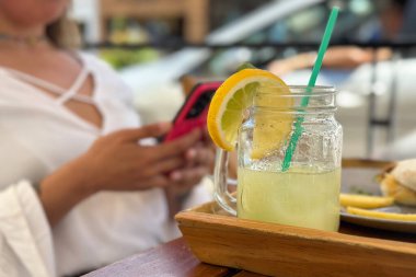 Arka planda cep telefonu kullanan tanınmayan bir kadınla kaldırımdaki kafede taze limonata masası.