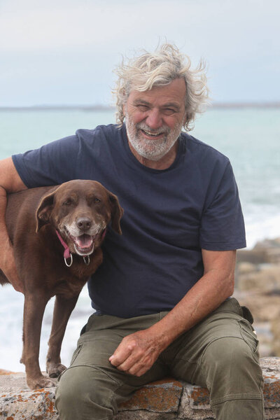 портрет старшего латиноамериканца, сближающегося со своей собакой на открытом воздухе с океаном на заднем плане