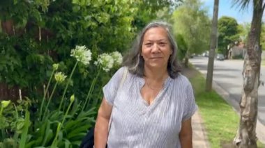 Yaz günü arka planda ağaçlar ve bitkilerle sokakta gülümseyen olgun Latin kadın portresi.