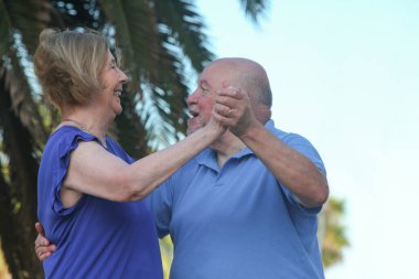 elderly man and woman dancing and smiling outdoors on summer day with palm trees and sky in background clipart