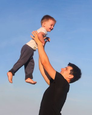 Genç baba, küçük oğlunu gün batımında güneşli gökyüzüne fırlatırken yukarıda tutuyor..