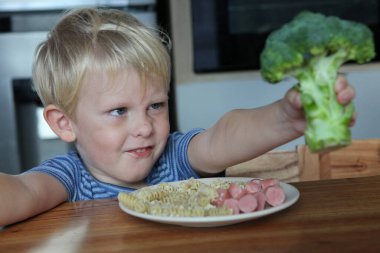 Tabakta erişte ve sosisle oturan şirin bir bebeğin portresi iğrenç bir suratla brokoliyi reddediyor.