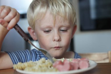 Gülümseyen bebek evde sosis ve erişte tabağıyla masada oturuyor.
