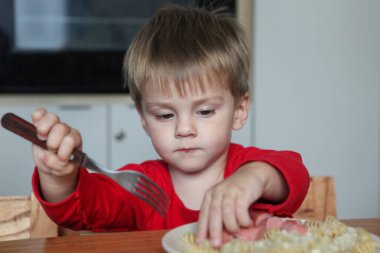 Gülümseyen çocuk elinde erişte ve sosisle masada oturmuş çatal-mutlu yemek zamanını evde kullanmayı öğreniyor.
