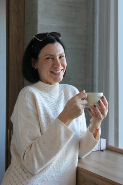 50 'li yaşlarının ortasında yetişkin bir kadın pencerenin önünde durmuş, kahve ya da çay içiyor, sessizliğin tadını çıkarıyor..